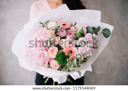 Very nice young woman holding big beautiful blossoming bouquet of fresh hydrangea, roses, barbara, matthiola, eucalyptus flowers in tender pink colors on the grey wall background