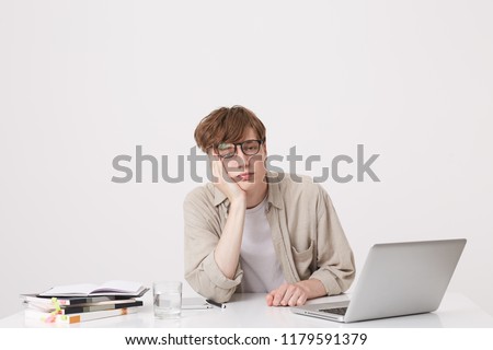 Portrait of sad bored young man student wears beige shirt and spectacles looks tired sitting at the table with laptop computer and notebooks isolated over white background