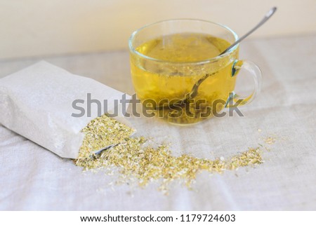 white herbal tea with dried chamomile flowers, tea in glass cup with spoon near white paper bag and dry scattered camomile, stock photo image