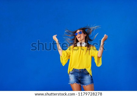 Cheerful girl in yellow shirt standing against blue wall with headset around her neck. Dancing and smiling.