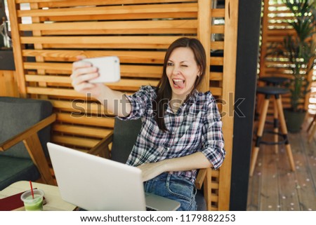 Woman in wooden outdoors street summer coffee shop sitting with laptop pc computer, doing selfie shot on mobile phone, relaxing during free time. Mobile Office. Lifestyle freelance business concept