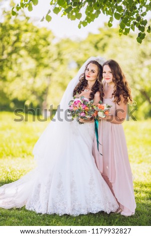 Young bride and her bridesmaid hugging in beautiful summer park. Best friends. Next bride wedding. Girls hug ech other before wedding ceremony. Cozy summer picture.