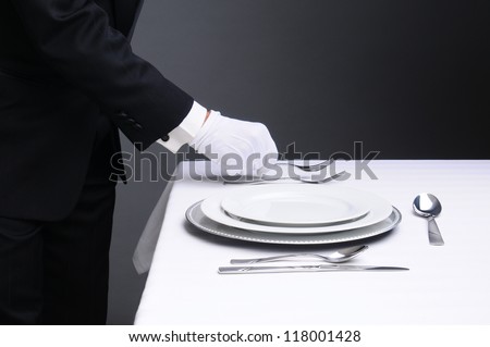 Closeup of a waiter in a tuxedo setting a formal dinner table. Horizontal format on a light to dark gray background. Man is unrecognizable. Royalty-Free Stock Photo #118001428