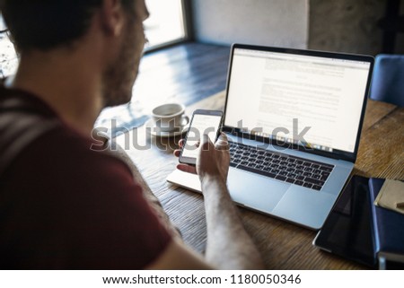Man professional content writer using apps on contemporary cellphone during distance job on laptop computer. Male freelancer online booking via cell telephone during remote job on portable netbook 