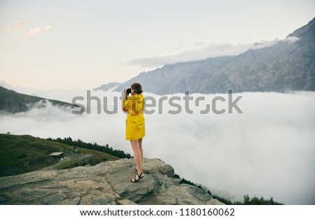 A young Girl in a yellow raincoat tourist photographs the mountains. . Girl making a photo shoot of mountain. Georgia. Summer. August. Mount Kazbegi.