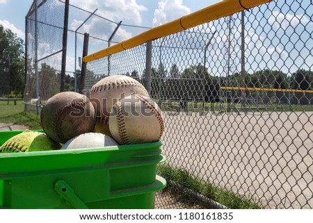 bucket with baseballs and softballs with field as background in daylight