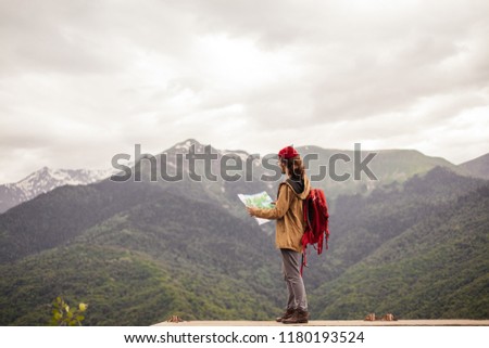 Young Man Traveler with map and red backpack searching location outdoor with rocky mountains on background