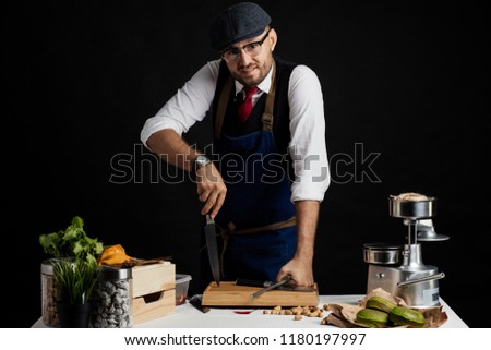 Restaurant Owner in cap and blue apron leaning on big Chief Knife in the kitchen near the wooden table against black background.