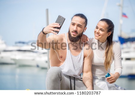 Resting after workout young cheerful caucasian couple in sports outfit taking selfie over seascape, blurred pier with ships, with smartphone.