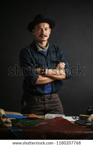 Portrait of handsome Leather goods craftsman in stylish hat doing handbags and leather accessories for ladies in his workshop on black dark background. Small business concept photo.