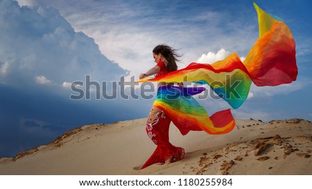
Belly dancer in a red dress, suit is dancing an oriental, East dance in the desert on the sand. Girl is holding rainbow shawl.