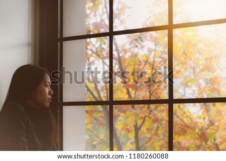 Asian beautiful woman thinking  and standing in room with autumn leaf outside window background.Concept of foreign students homesick.