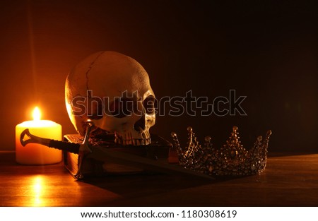 Human skull, old book, sword, crown and burning candle over old wooden table and dark background