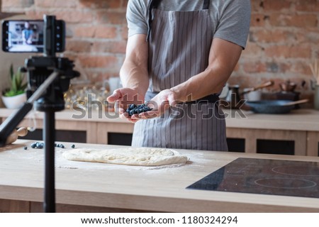 social media influencer or food blogger creating content. man shooting a cooking video using camera on tripod. chef holding bluberries in hands and showing them to viewer. Royalty-Free Stock Photo #1180324294
