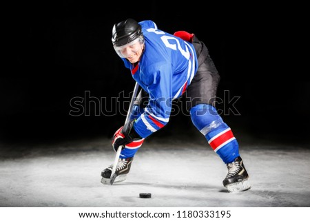 Young hockey player with stick and puck skating on rink in attack against dark background