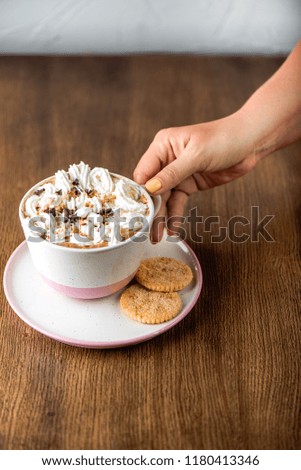 Coffee with marshmallows, cream, grated chocolate, nuts in large cup. Woomen hand holds a cup. A beautiful serving of dessert, cookies in the frame, a wooden table. Place for text