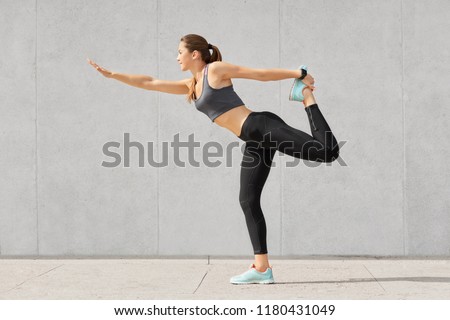 Attractive slim woman enjoys active lifestyle, makes warming up exercises before starting workout session, dressed in sportswear, shows her flexibility, sporty body, poses against grey wall.