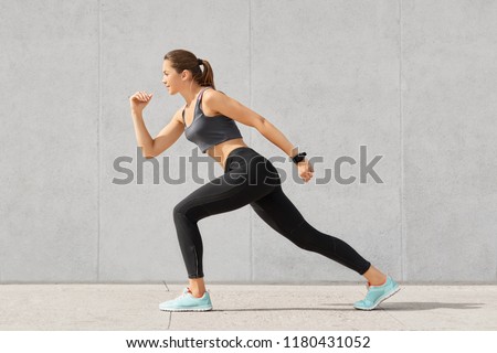 Horizontal shot of flexible woman with sporty body, stands in pose, ready to start marathon, dressed in sportsclothes, isolated over grey concrete wall. People. sport, determination concept.