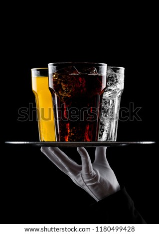 Hand with glove holds tray with glasses of colas soda drink, orange and water on black background with ice cubes