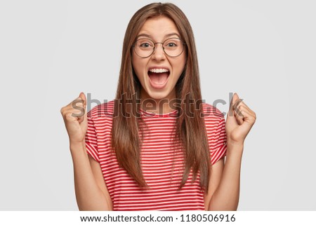 Self determined woman with dark straight hair, clenches fists and exclaims with triumph expression, cheers about something good, screams: Yes, I did it finally! People and body language concept Royalty-Free Stock Photo #1180506916