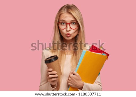 Lovely blonde schoolgirl keeps lips rounded, flirts with handsome guy at school, wears round spectacles with red rims for good vision, holds colourful textbooks, disposable cup of coffee or cappuccino