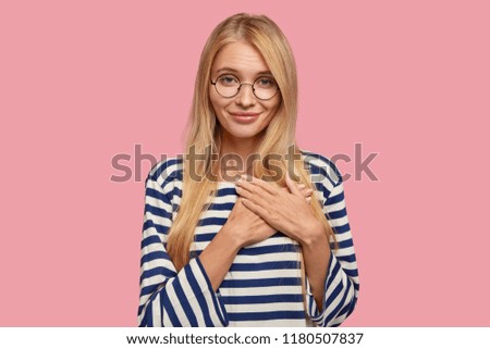Cheerful pleasant looking girl keeps hands on chest, looks with love and tender attitude, stops breathing from pleasant feelings, expresses gratitude, dressed casually, isolated on pink wall