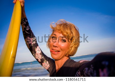 young attractive and happy blonde surfer woman in swimsuit holding surf board in the beach taking self portrait selfie picture smiling playful enjoying holidays at tropical island