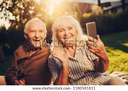 Hilarious laughing mature pair sitting on grass and looking at handy in female hand with bright smile. Portrait