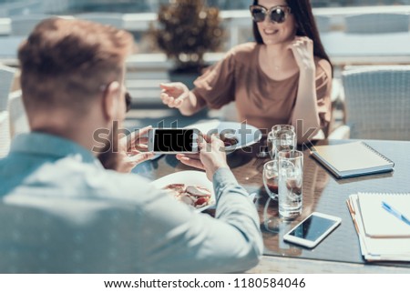 Male talking photo of dish while looking at screen of phone. Positive female talking with him while gesticulating hand. They sitting at desk in cafe during conversation