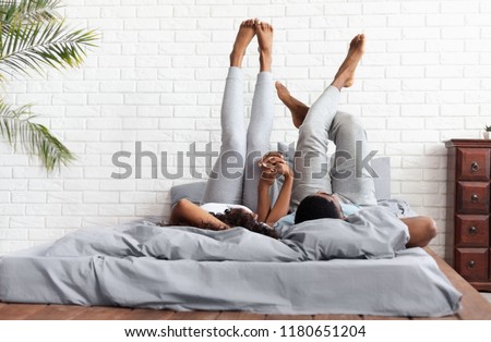 Couple in love. Young happy african-american man and woman lying on bed at home raising legs up, lazy family resting on weekend, copy space Royalty-Free Stock Photo #1180651204