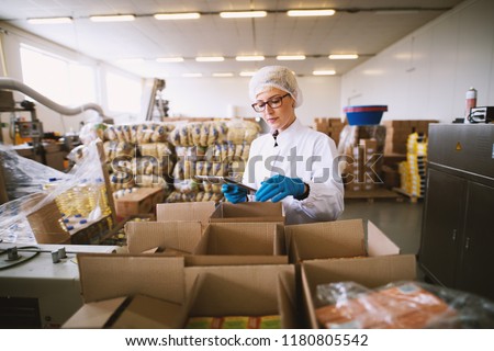 Young female worker in sterile clothes is using tablet to check right number of packages.