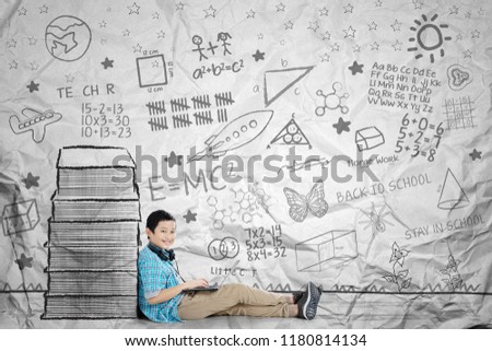 Picture of Asian preteen schoolboy using a laptop while sitting near a pile of drawn books. Shot with crumpled background
