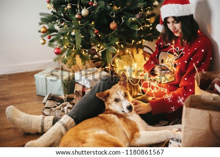 happy girl in santa hat holding phone and hugging dog under golden beautiful christmas tree with lights and presents in festive room. winter atmospheric  moments. happy holidays