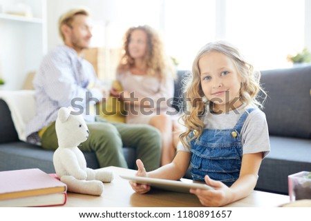 Little girl with tablet looking at you while watching videos at home with her parents on background