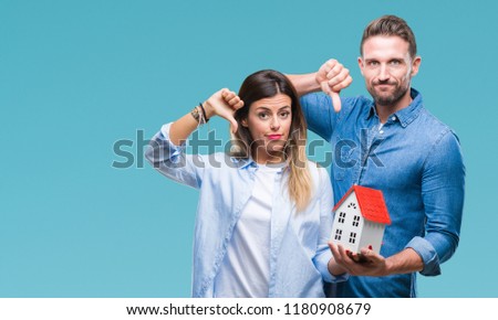 Young couple in love holding house over isolated background with angry face, negative sign showing dislike with thumbs down, rejection concept