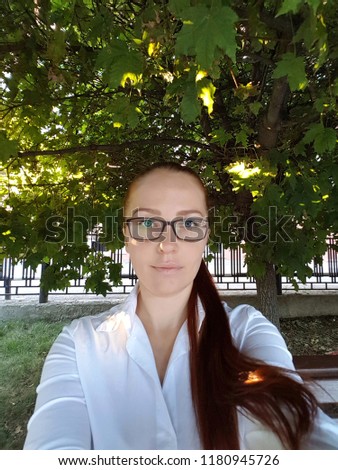 a beautiful business Caucasian middle-aged woman with red hair in optical glasses sits on a bench in the Park and takes a selfie on her smartphone.