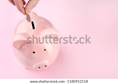 Female hand putting coin into piggy bank on pink background
