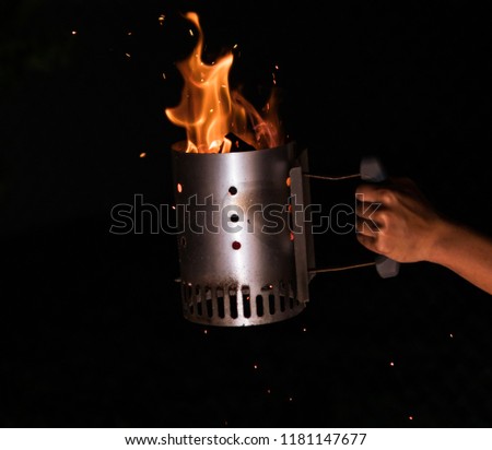 Photo of a grill / chimney starter with large flames and sparks coming off of it being held by a hand during the evening.