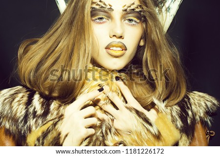 Closeup portrait of one beautiful wild young woman with bright golden animal monkey makeup with thorns on face and antlers in fur coat on black background, horizontal picture