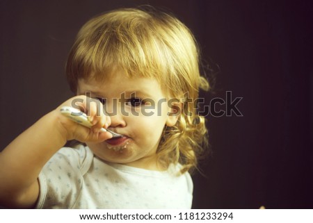 Closeup portrait view of one adorable cute small baby boy with blonde hair eating healthy food of porridge or coocked semolina with spoon in hand indoor, horizontal picture