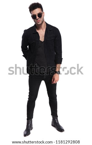 handsome macho man in black clothes and wearing sunglasses standing on white background with a hand behind his back, full length picture