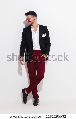 attractive smart casual man looks to side and smiles while leaning against a white wall, looking relaxed, full body picture