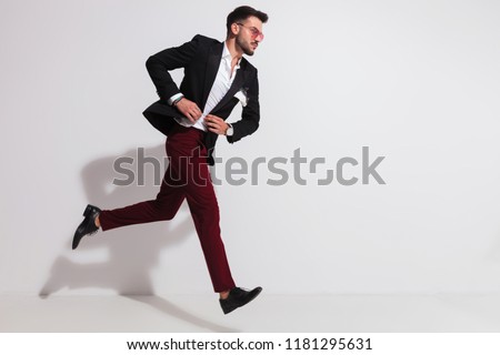side view of elegant man with red sungless running near a white wall and buttoning his black suit, full length picture