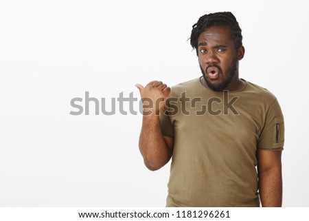 Questioned unsure attractive dark-skinned male coworker in military t-shirt, pointing backwards or left with thumb, asking question with doubtful intrigued expression, standing over grey wall