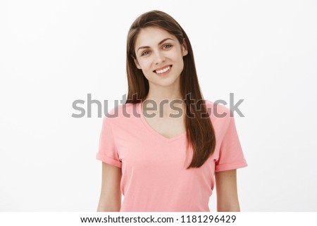 Friendly good-looking female coworker in pink t-shirt smiling broadly and tilting head, listening or talking with customer, being polite and in good mood while standing against gray background