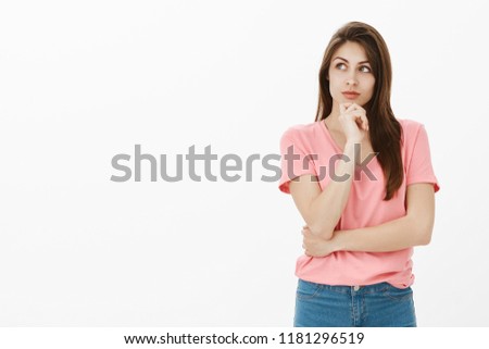 Stylish and smart businesswoman thinking about getting day off. Portrait of good-looking dreamy girlfriend in pink t-shirt, holding hand on chin, looking left with interest while picturing in mind
