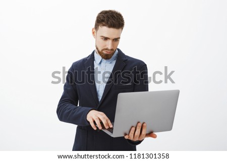 Focusing on business. Portrait of smart and ambitious concentrated good-looking young male entrepreneur with beard and blue eyes holding laptop in hand browsing checking schedule with determined look