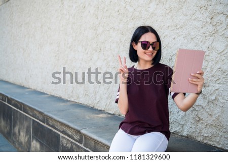 Self portrait of cheerful positive girl shooting selfie on front camera with two hands having video-call with friend gesturing v-sign outdoors