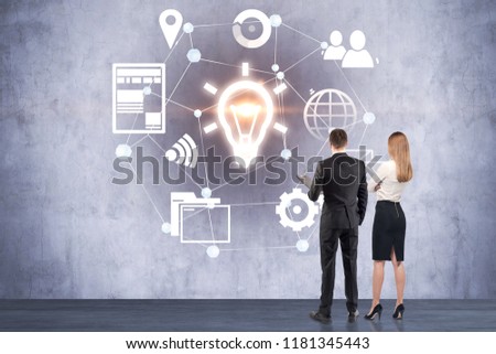 Businesswoman and businessman looking at glowing management and business idea icons drawn on a concrete wall. Talented leader concept. Toned image copy space