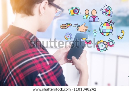 Hipster geek girl wearing glasses and checkered shirt holding black screen smartphone in office. Bright and colorful hand drawn social media icons and texts above it. Toned image double exposure